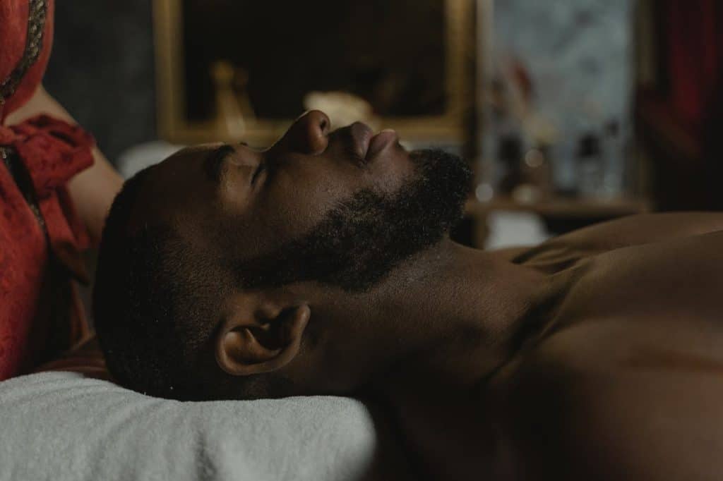Homem negro deitado de bruços em uma maca de massagem, com os olhos fechados e expressão relaxada, recebendo uma massagem, ilustrando a experiência de uma "massagem em Natal" focada em bem-estar.