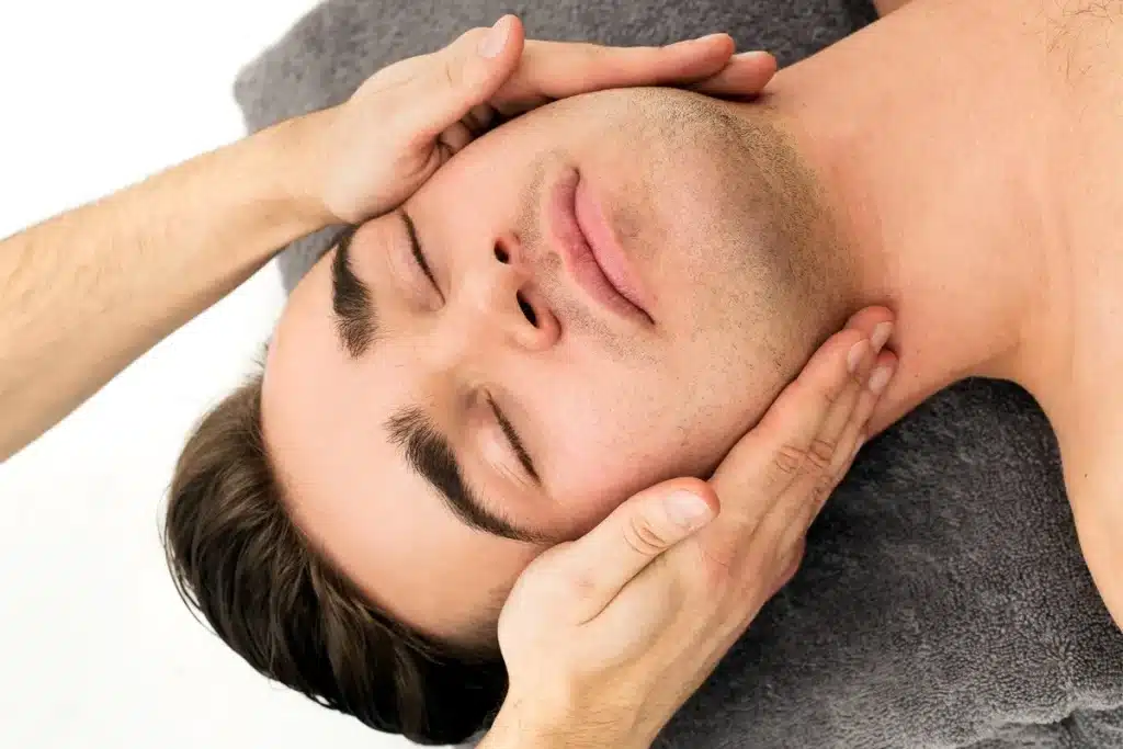 Homem relaxando com os olhos fechados enquanto recebe massagem no rosto, com as mãos do terapeuta aplicando pressão suave nas laterais da cabeça.