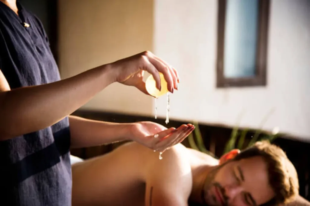Close-up de massagista profissional derramando óleo de massagem (óleo essencial ou terapêutico) nas mãos, em preparação para iniciar uma sessão em um cliente deitado de bruços. A cena de massagem em Niterói transmite relaxamento e cuidado.