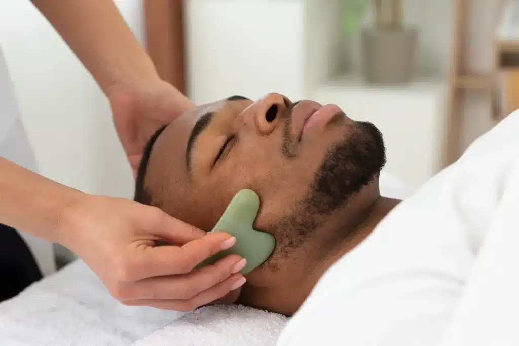 Terapeuta realizando massagem facial relaxante com pedra Gua Sha em um homem, promovendo relaxamento e bem-estar.