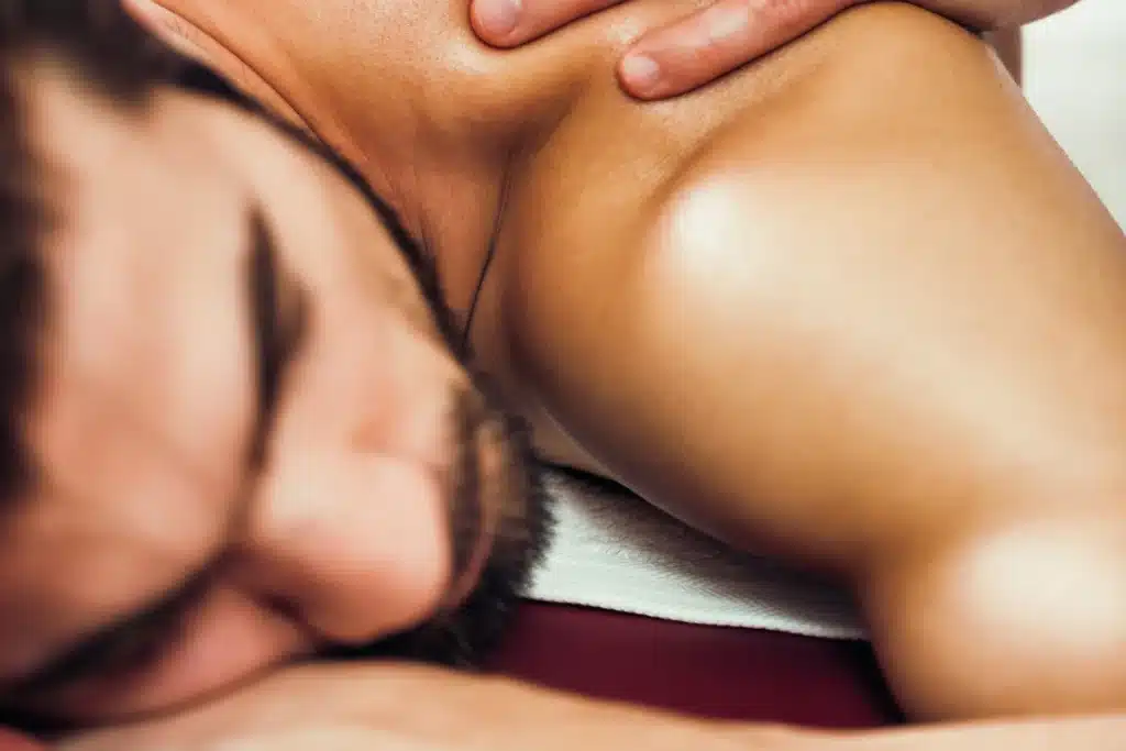  Close-up de um homem recebendo massagem no ombro com foco na pele e nas mãos do terapeuta.