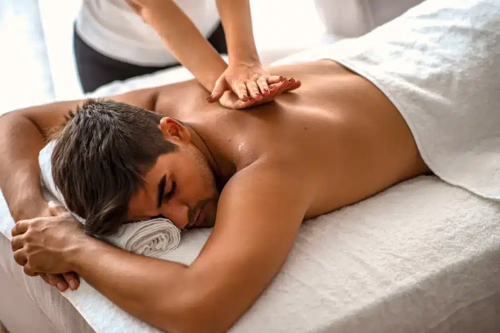 Homem recebendo massagem relaxante em spa, deitado sobre uma maca com toalha branca, enquanto profissional aplica técnica manual nas costas.