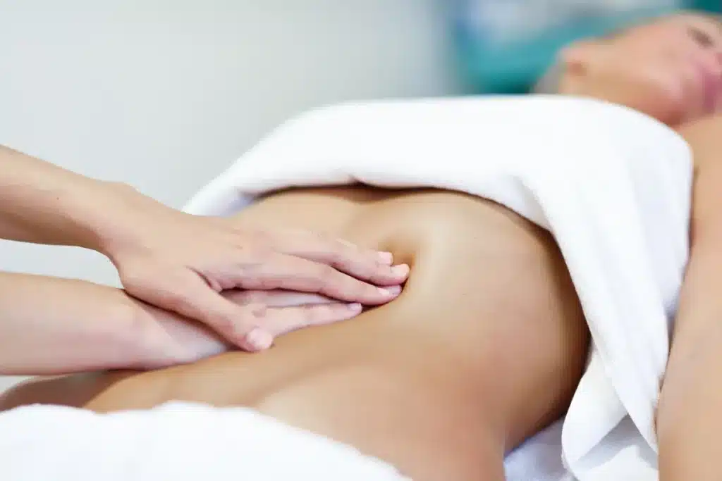 Mãos aplicando uma massagem relaxante na barriga de uma pessoa deitada, coberta com uma toalha branca.