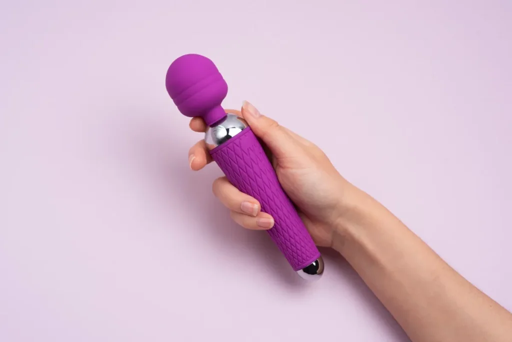 Mão segurando um massageador pessoal vibratório roxo com design ergonômico sobre um fundo lilás.