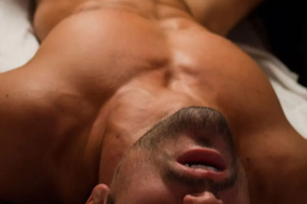 Close-up de um homem deitado em uma maca de massagem, com o peito e o pescoço em destaque, os olhos fechados e a boca entreaberta, sugerindo um momento de relaxamento intenso, êxtase ou prazer durante uma sessão de massagem com finalização. A iluminação suave foca na pele bronzeada e nos músculos do peito.