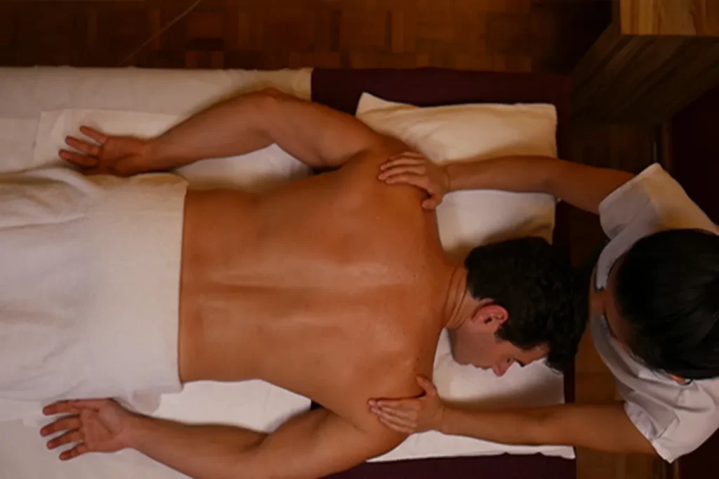 Vista de cima de uma massagista aplicando massagem masculina nas costas e ombros de um cliente deitado de bruços, coberto por toalha branca.