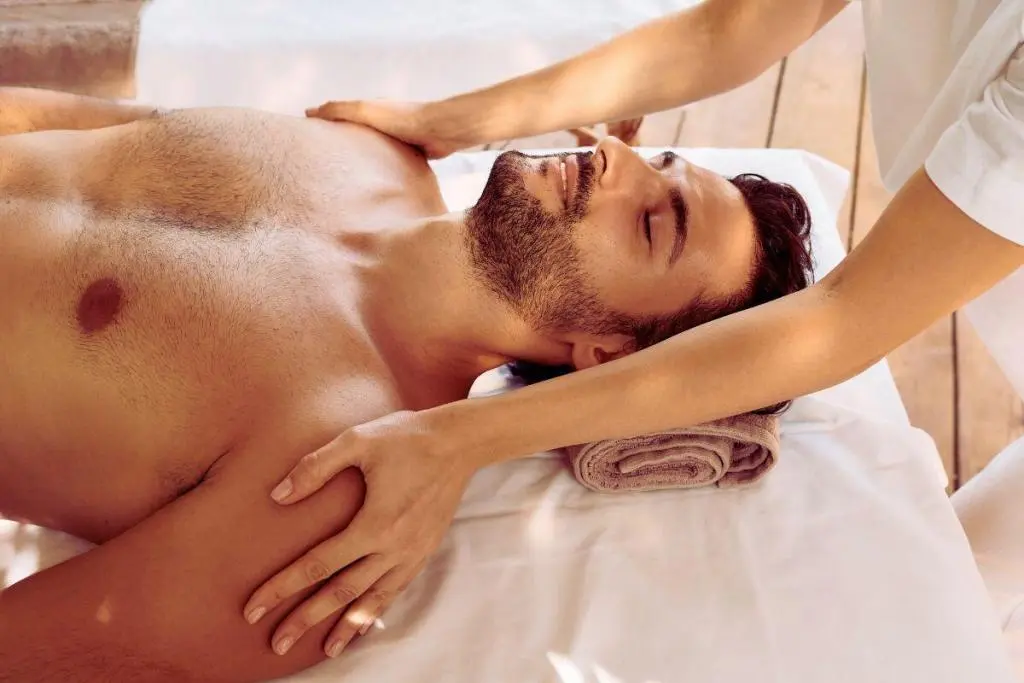 Homem deitado recebendo massagem relaxante nos ombros e peito, com expressão serena em ambiente tranquilo.