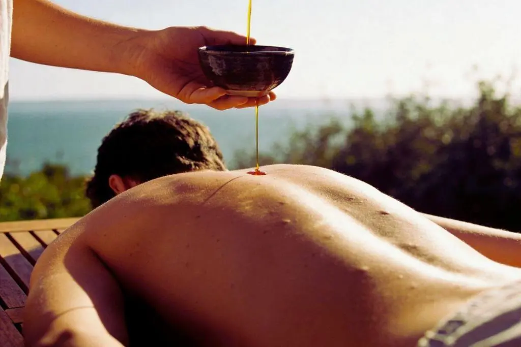 Aplicação de óleo terapêutico nas costas de uma pessoa durante sessão de massagem relaxante ao ar livre com vista natural
