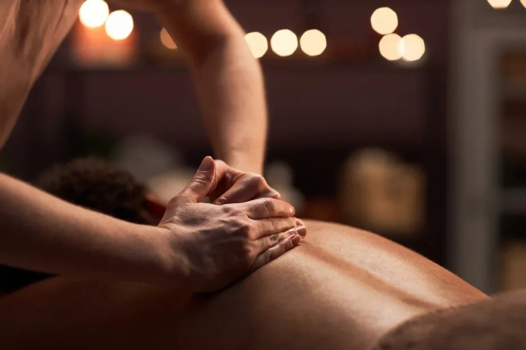 mãos realizando massagem nas costas de um homem em ambiente com luz baixa e velas ao fundo