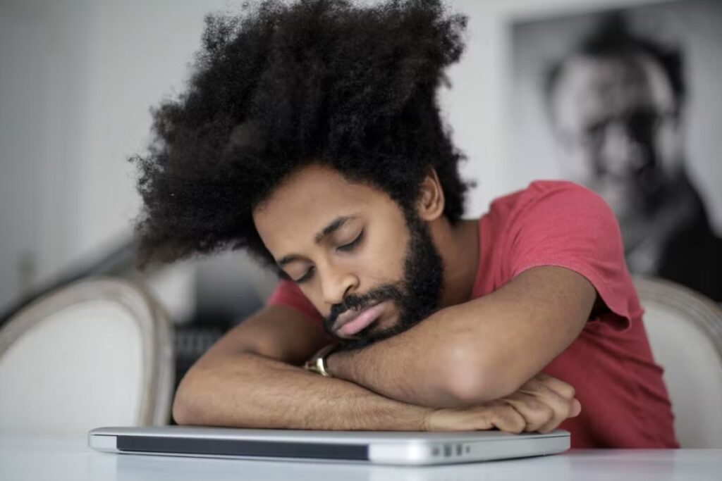 Homem negro de camiseta vermelha com a cabeça apoiada sobre os braços cruzados em cima de um notebook fechado, aparentando dormir ou descansar.