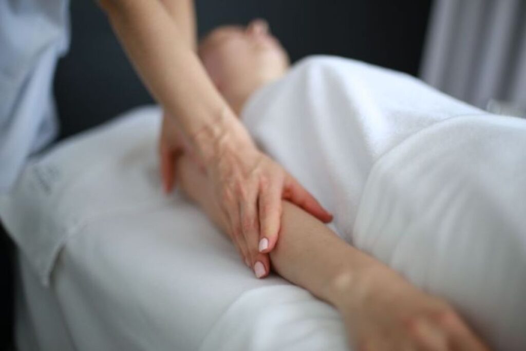 Close de mãos aplicando uma massagem suave no braço de uma pessoa coberta por um lençol branco.