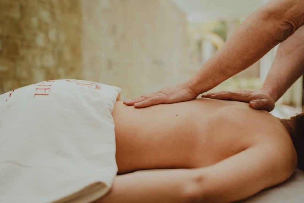Vista lateral aproximada das costas de uma pessoa deitada de bruços, recebendo massagem terapêutica com as duas mãos do profissional apoiadas na região lombar e média.