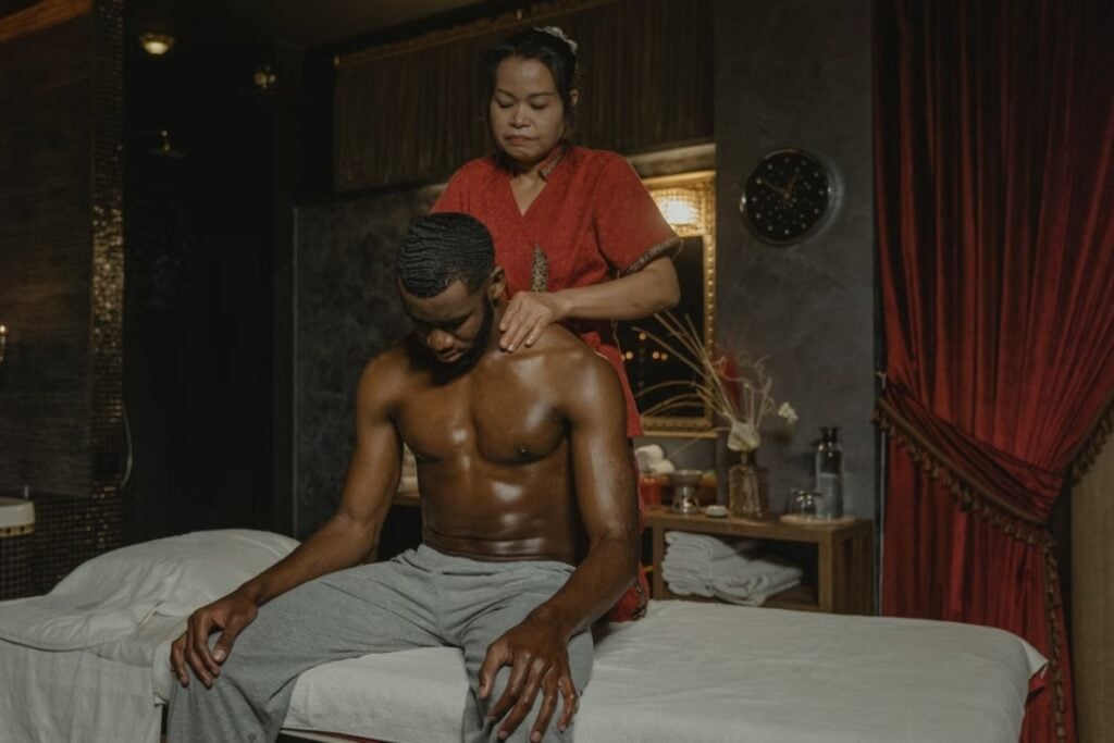 Homem negro sentado em uma maca recebendo massagem nos ombros por uma profissional vestida de vermelho em um ambiente com cortinas escuras.