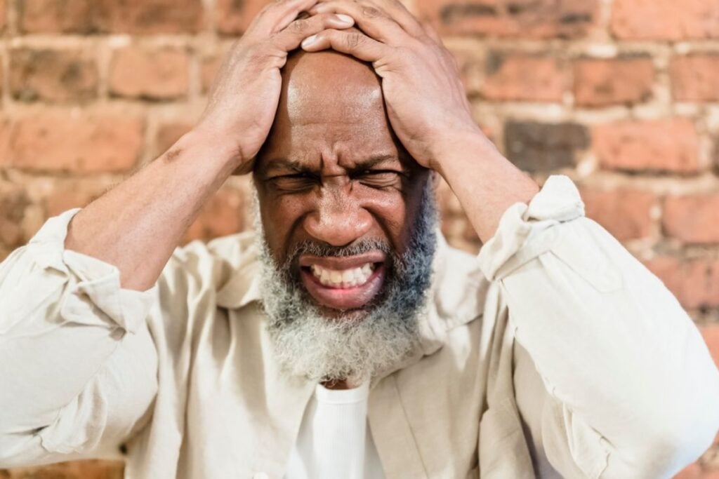 Homem idoso negro, de barba grisalha, com as duas mãos espalmadas nas laterais da cabeça, olhos fechados e dentes cerrados diante de uma parede de tijolos.