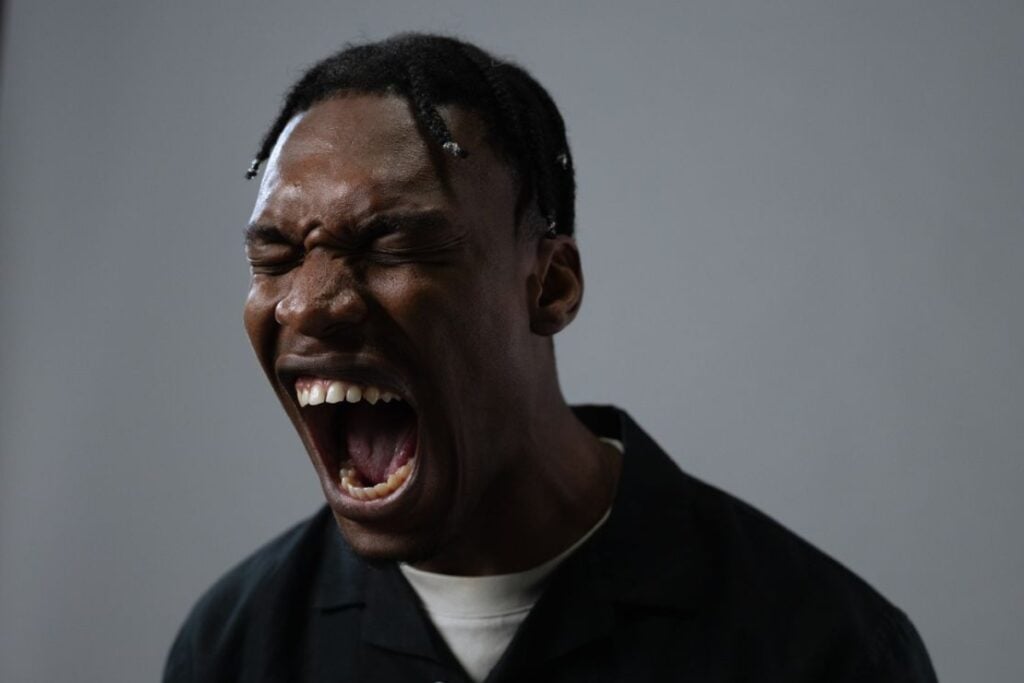 ovem negro de perfil parcial com os olhos bem fechados e a boca totalmente aberta em expressão de grito, contra um fundo cinza.