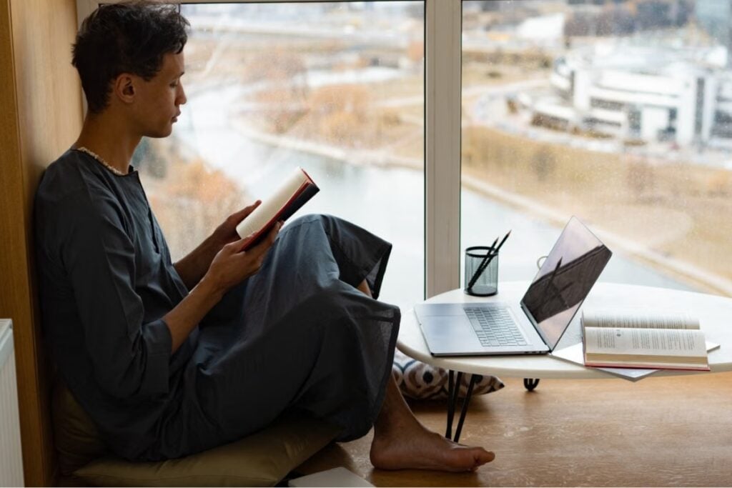 Homem sentado de perfil no chão com as pernas cruzadas, segurando um livro aberto em frente a uma grande janela de vidro, ao lado de uma mesa com notebook.