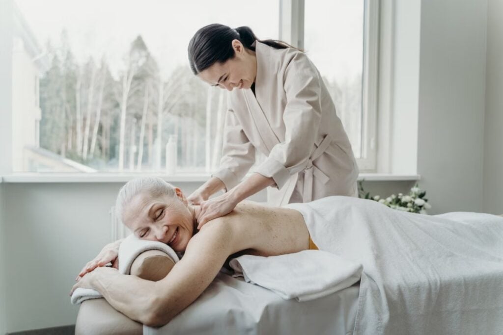 Mulher idosa deitada de bruços sorrindo enquanto recebe uma massagem nos ombros de uma terapeuta em um ambiente iluminado.