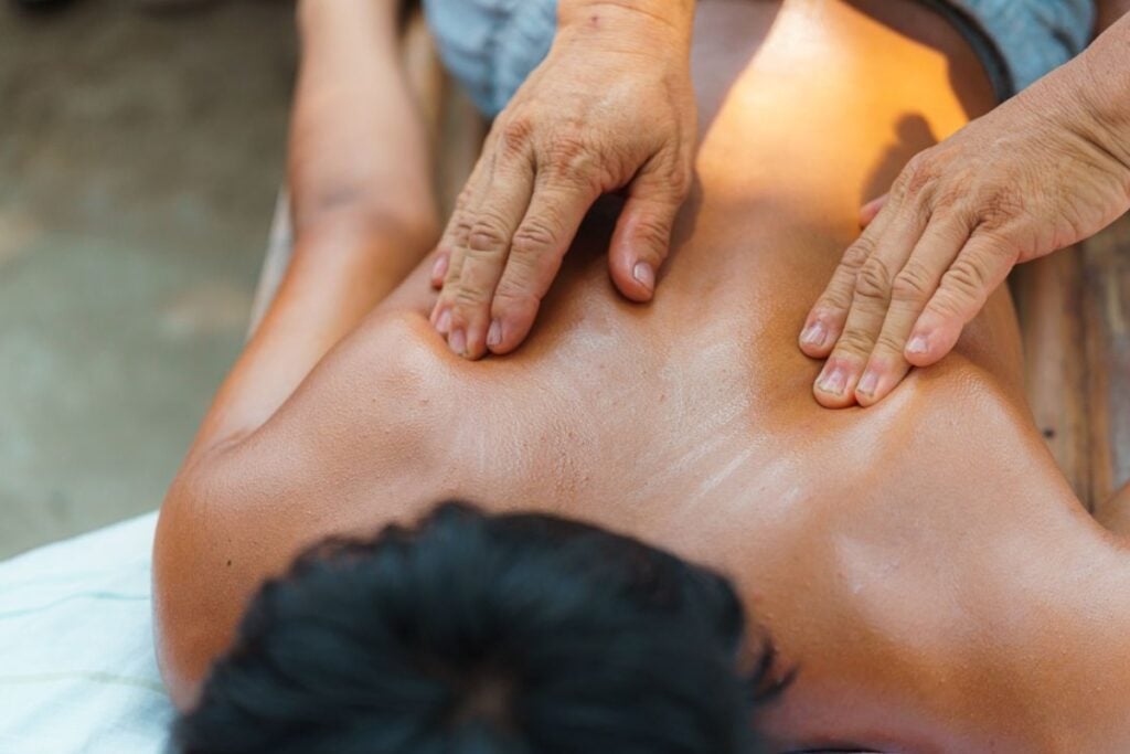 Vista superior em close das costas de um homem recebendo massagem com as duas mãos de um terapeuta aplicando pressão nos ombros.
