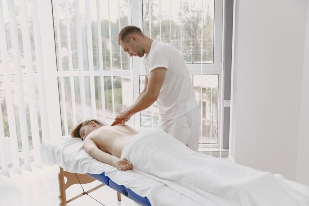 Terapeuta de camiseta branca aplicando massagem nas costas de uma mulher deitada em uma maca próxima a uma grande janela com persianas verticais.