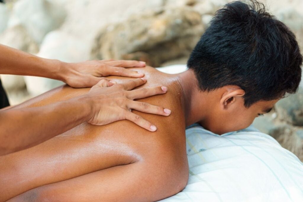 Vista lateral aproximada de um jovem deitado de bruços recebendo massagem nas costas com as duas mãos do terapeuta posicionadas sobre a região superior do tronco.