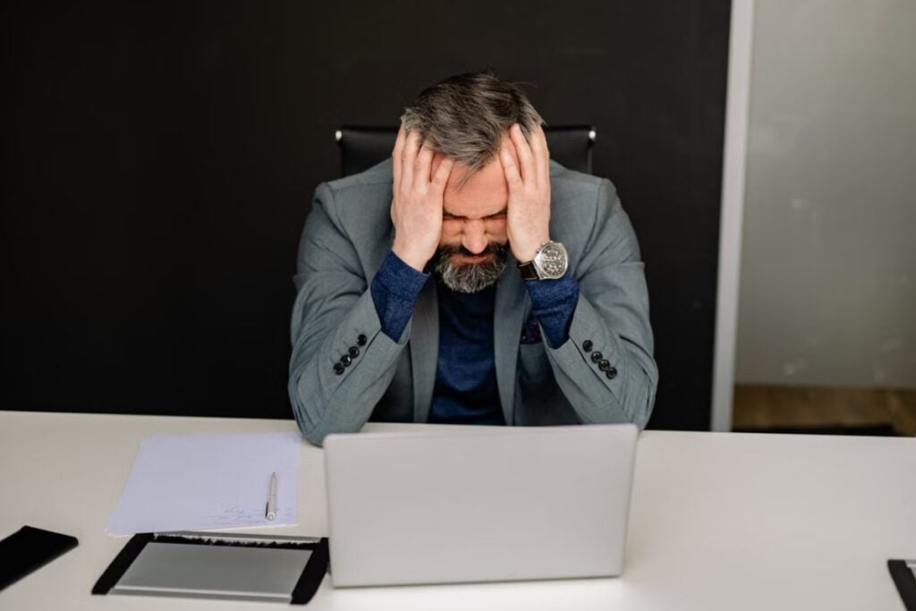 Fotografia de plano médio mostrando um homem de meia-idade, com barba grisalha e terno cinza, sentado à mesa de trabalho com as mãos nas têmporas e os olhos fechados em uma expressão de forte dor de cabeça ou estresse, diante de um notebook aberto.