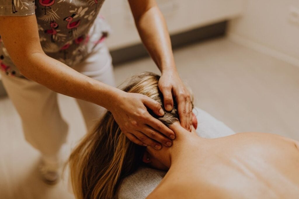 Fotografia em plano detalhe e ângulo superior mostrando as mãos de uma terapeuta pressionando e massageando a nuca e a base do crânio de uma mulher loira deitada de bruços em uma maca de massagem, em um ambiente clínico iluminado.
